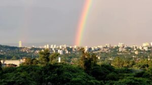 Arco-íris iluminando a paisagem urbana com prédios e mata ao entardecer, imagem que inspira a escrita de um haicai urbano.